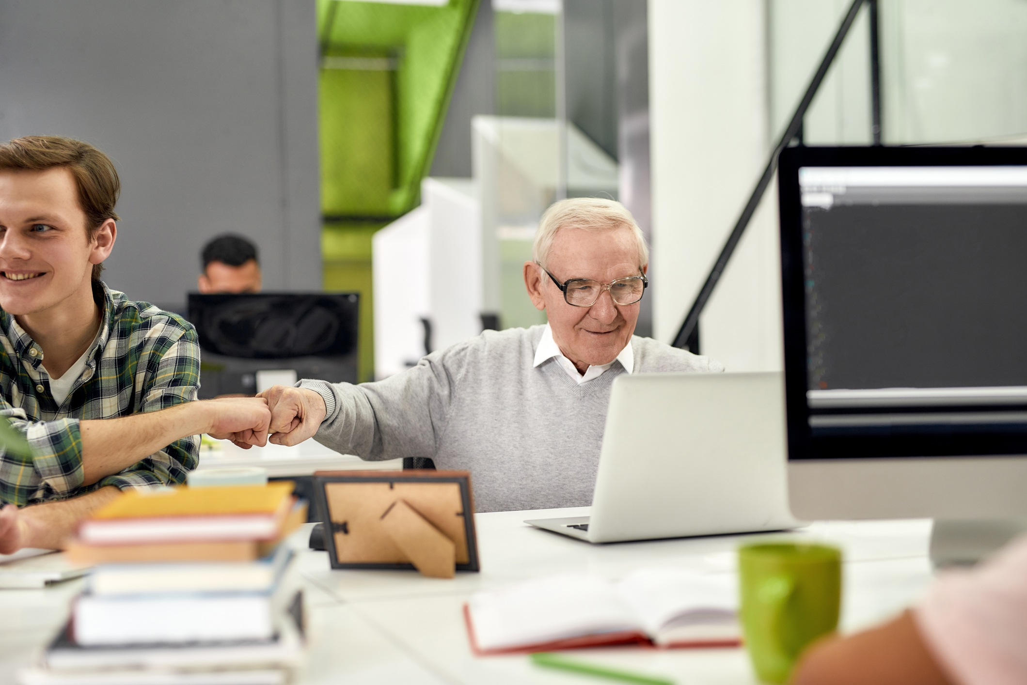 Friendly. Aged man, senior intern looking at the screen of his laptop and doing a fist bump with colleague